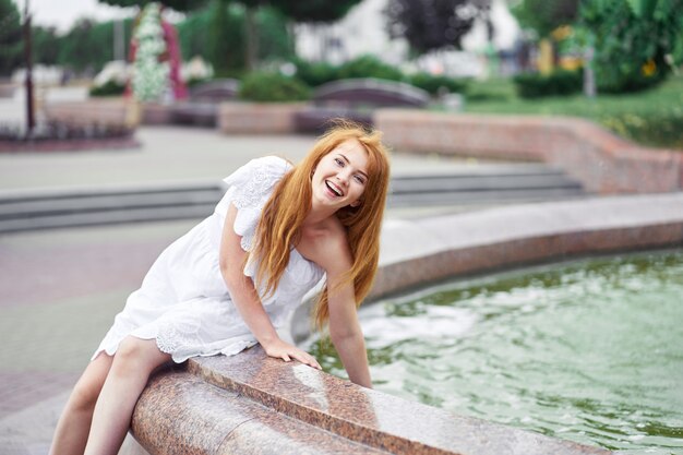 Ragazza felice attraente alla moda della testarossa vicino alla fontana nella città.