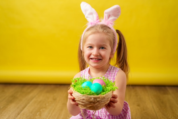 Ragazza felice bionda del bambino con le orecchie del coniglietto e canestro delle uova per Pasqua