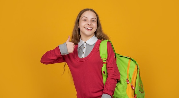 Ragazza felice che dà il pollice che porta la borsa della scuola sfondo giallo 1 settembre