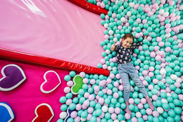 Ragazza felice che gioca al parco giochi del centro giochi al coperto Sdraiato sulle palle di colore nella piscina di palline