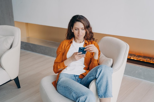 Ragazza felice che si siede e manda messaggi sul cellulare a casa e ascolta musica con gli auricolari wireless