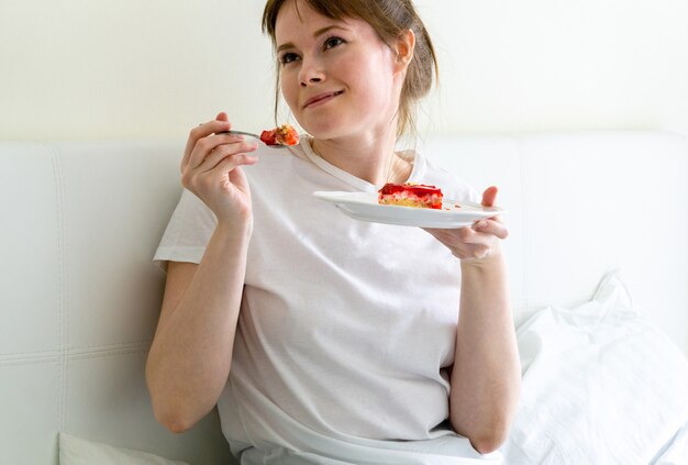 Ragazza felice che si siede sul letto e che mangia dolce