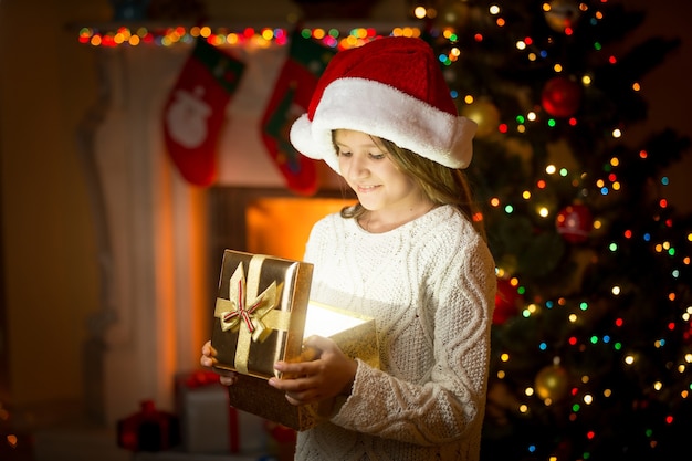 Ragazza felice con cappello da Babbo Natale che tiene in mano una scatola regalo incandescente