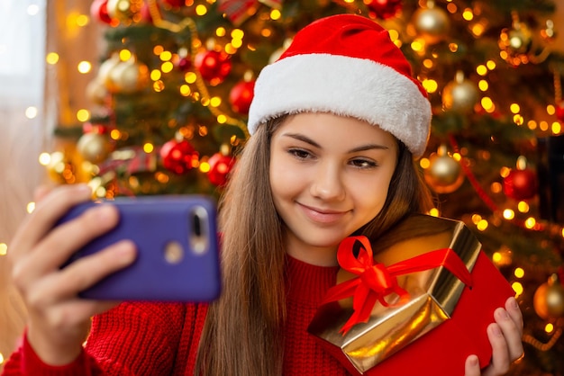 Ragazza felice con cappello da Babbo Natale e scatola rossa nelle sue mani che fanno selfie Vacanze invernali di Natale Capodanno ottenendo regali concetto