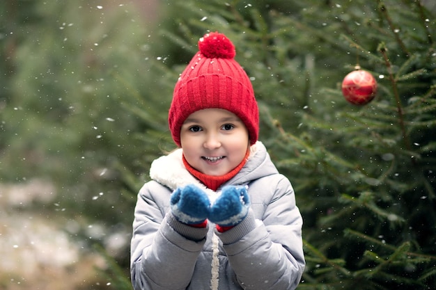 Ragazza felice con il berretto rosso che gioca all'aperto nella foresta invernale innevata e soffiando la neve