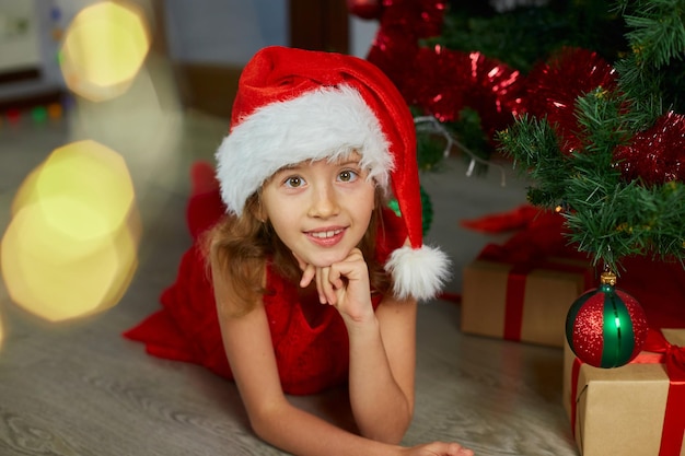 Ragazza felice con il cappello di Babbo Natale che si trova vicino all'albero di Natale a casa