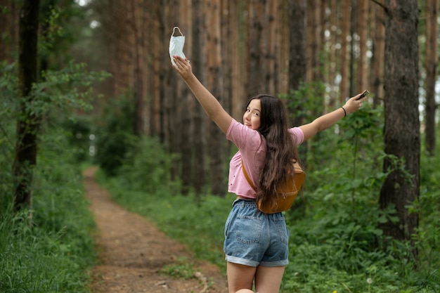 Ragazza felice con la maglietta rosa e lo zaino dietro la schiena che cammina nella foresta. La ragazza senza mascherina medica gode dell'aria fresca e pulita.