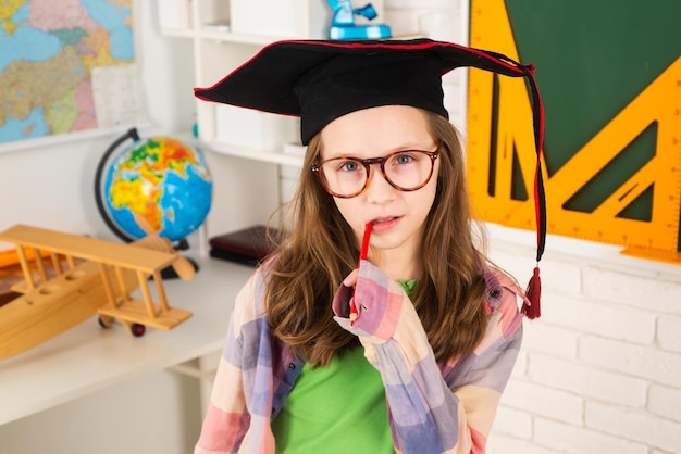 Ragazza felice con la penna in bocca nel cappello di laurea vicino a un consiglio scolastico