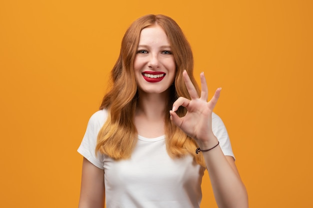 Ragazza felice con la testarossa ondulata lunga, maglietta bianca da portare sorridente e che mostra il gesto GIUSTO.