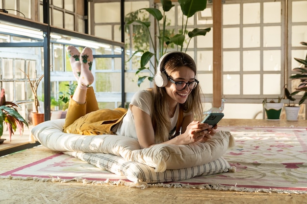 Ragazza felice con le cuffie che ascolta la musica sul suo telefono che si rilassa sdraiata sul pavimento a casa