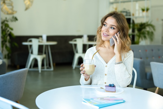 Ragazza felice con un bicchiere di cappuccino e dessert ai mirtilli parlando da smartphone mentre vi rilassate nella caffetteria