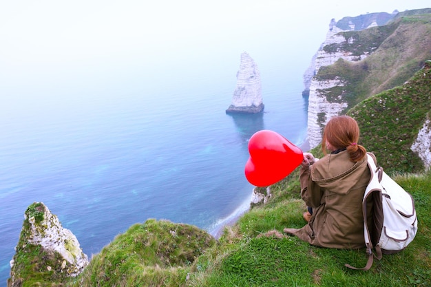 Ragazza felice con un palloncino rosso a forma di cuore sullo sfondo del paesaggio Etretat. costa settentrionale della Francia
