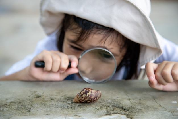 Ragazza felice del bambino che esplora la natura con una lente d'ingrandimento e una lumaca. Si diverte in giardino. Il concetto del bambino è pronto per andare a scuola.