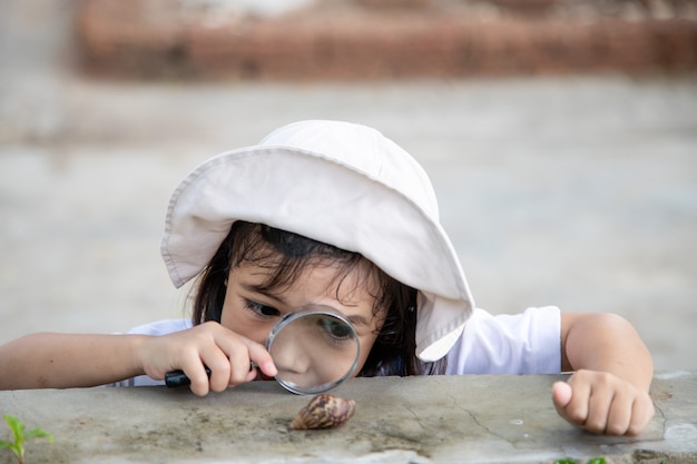 Ragazza felice del bambino che esplora la natura con una lente d'ingrandimento e una lumaca. Si diverte in giardino. Il concetto del bambino è pronto per andare a scuola.