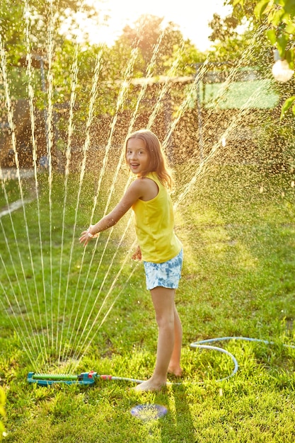 Ragazza felice del bambino che gioca con l'irrigatore da giardino correre e saltare l'estate