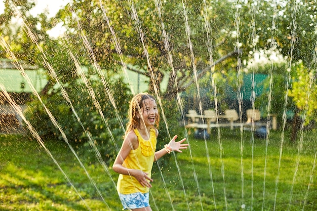 Ragazza felice del bambino che gioca con l'irrigatore da giardino correre e saltare l'estate