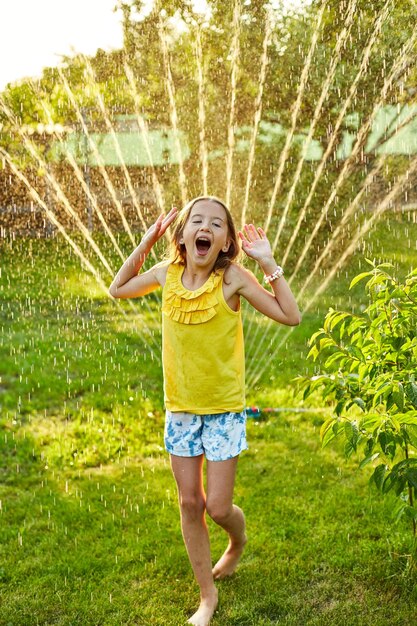 Ragazza felice del bambino che gioca con l'irrigatore da giardino correre e saltare l'estate