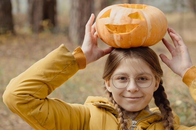 Ragazza felice del bambino con la zucca di Halloween sulla testa. Preparazione per la festa in giardino vicino alle decorazioni di Jack-o-Lantern