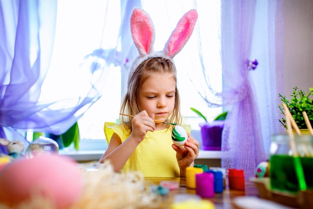 Ragazza felice di pasqua in orecchie del coniglietto che dipingono le uova, piccolo bambino a casa. vacanze di primavera