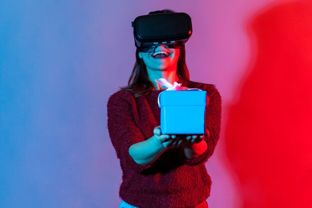 Ragazza felice e positiva in occhiali per realtà virtuale VR che tengono una confezione regalo avvolta e sorride gioiosamente