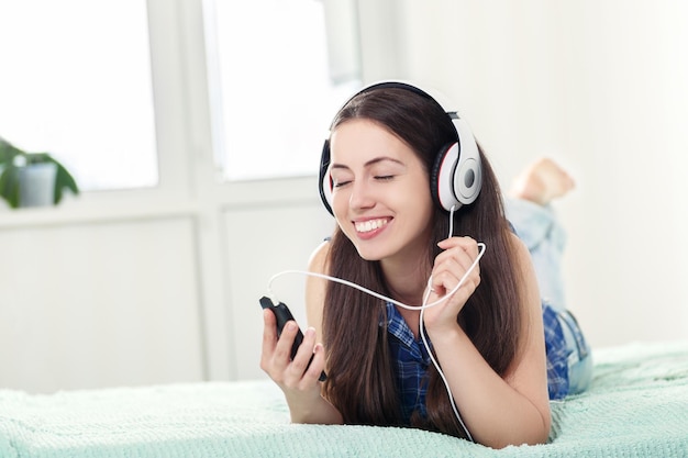 Ragazza felice e rilassata che ascolta musica con le cuffie e lo smartphone sdraiata sul letto a casa