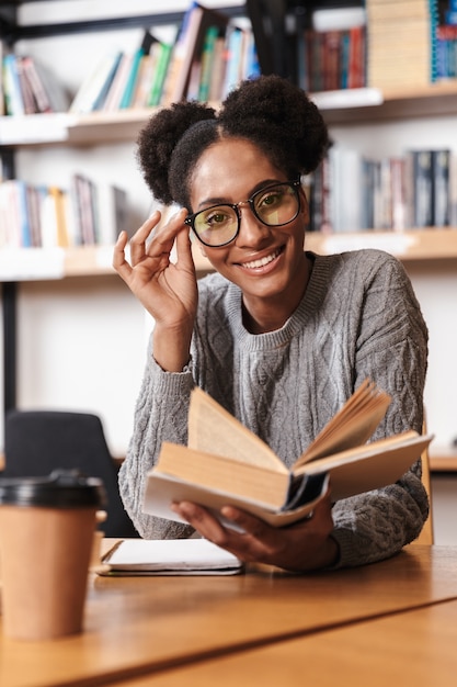 Ragazza felice giovane studente africano che studia in biblioteca, leggendo un libro