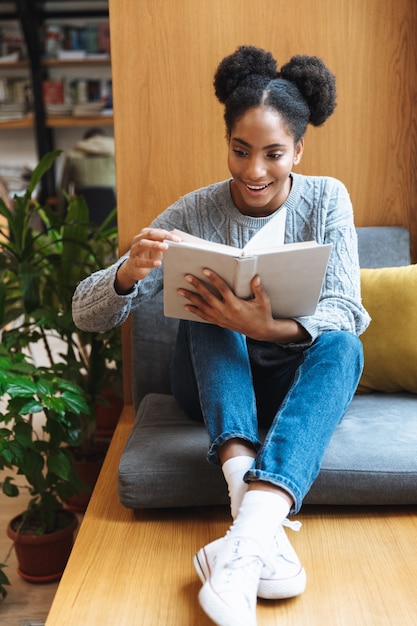 Ragazza felice giovane studente africano che studia in biblioteca, leggendo un libro