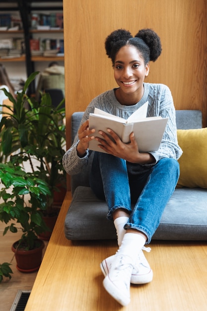 Ragazza felice giovane studente africano che studia in biblioteca, leggendo un libro