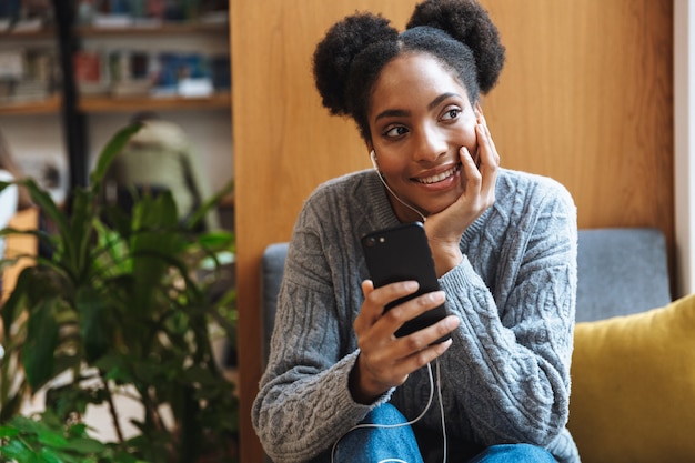 Ragazza felice giovane studente africano che studia presso la biblioteca, ascoltando musica con gli auricolari