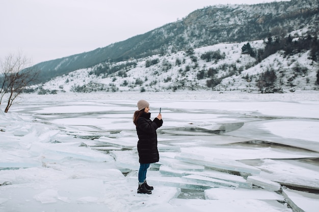 Ragazza felice in piedi su una riva del fiume in inverno e scattare foto al telefono. Ragazza che gode della vista del fiume ghiacciato.