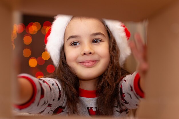 Ragazza felice in pigiama di Capodanno e cappello di Babbo Natale tira fuori un regalo dalla scatola