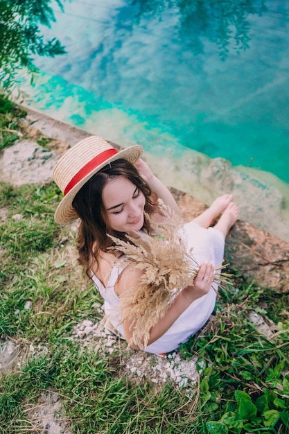 Ragazza felice in un vestito che si siede sulla riva di un lago blu. Ragazza in un cappello di paglia con un bouquet vicino a un piccolo lago.