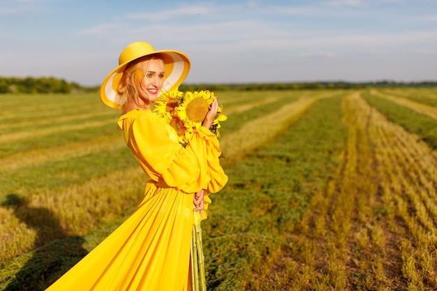 ragazza felice in un vestito giallo tiene fiori di girasole in un campo di girasoli contro il cielo