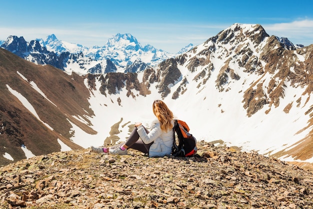 Ragazza felice in una giacca bianca con uno zaino seduto sul bordo di un picco di montagna. paesaggio di neve di montagna