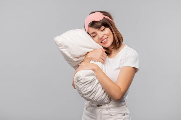 Ragazza felice nella maschera per dormire mantenendo la testa sul cuscino mentre sonnecchia in isolamento