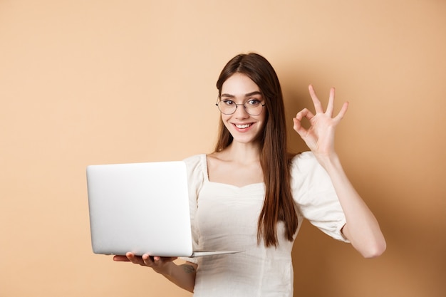 Ragazza freelance con gli occhiali che mostra il segno giusto e lavora al laptop, garantisce una buona qualità, consiglia qualcosa online, in piedi su sfondo beige.