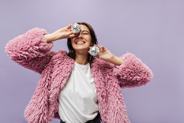 Ragazza gioiosa con capelli castani in maglione rosa caldo a maniche lunghe e maglietta bianca sorridente e in possesso di palle da discoteca su sfondo lilla