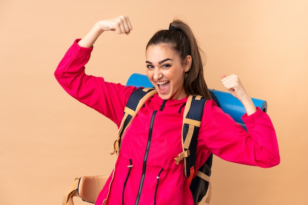 Ragazza giovane alpinista con un grande viaggiatore con zaino e sacco a pelo su beige che celebra una vittoria