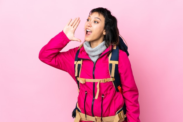 Ragazza giovane alpinista con un grande zaino sopra la parete rosa che grida con la bocca spalancata