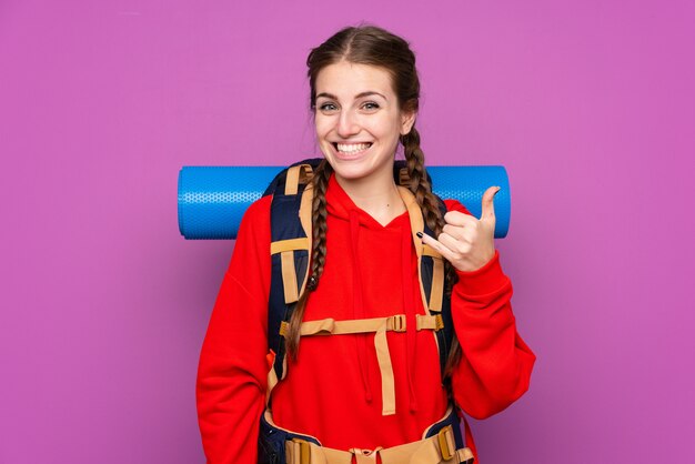 Ragazza giovane alpinista con un grande zaino sopra la parete viola isolata che fa gesto del telefono