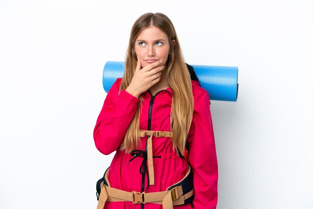 Ragazza giovane alpinista con un grande zaino su sfondo bianco isolato che ha dubbi e con un'espressione facciale confusa