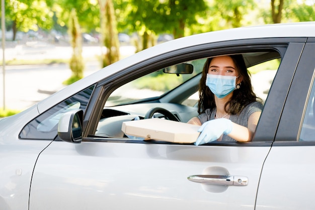 Ragazza giovane con scatola pizza in auto con mascherina medica e guanti