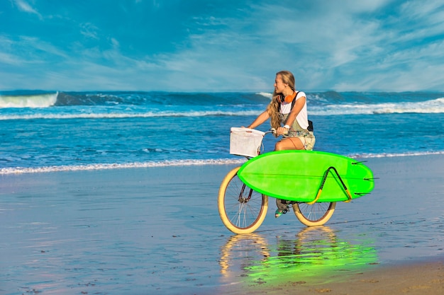Ragazza giovane con tavola da surf e bicicletta sulla spiaggia