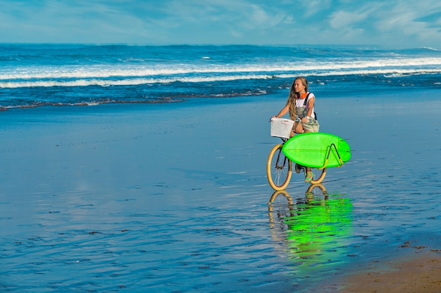 Ragazza giovane con tavola da surf e bicicletta sulla spiaggia