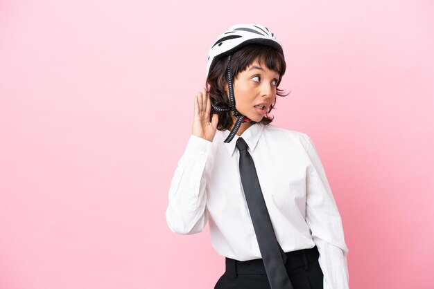 Ragazza giovane con un casco da bici isolato su sfondo rosa che ascolta qualcosa mettendo la mano sull'orecchio