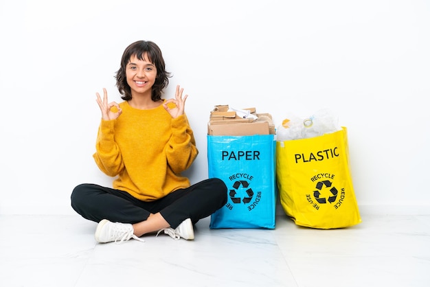 Ragazza giovane con una borsa piena di plastica e carta seduta sul pavimento isolato su sfondo bianco che mostra un segno ok con le dita