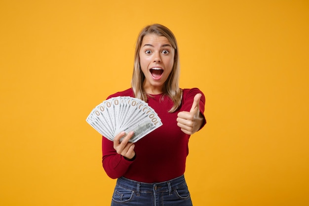 Ragazza giovane donna bionda stupita in abiti casual in posa isolato su sfondo giallo arancione. Concetto di stile di vita delle persone. Mock up copy space Tenere il ventaglio di denaro contante in banconote in dollari che mostrano il pollice in alto.