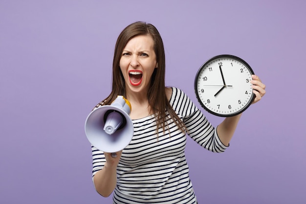 Ragazza giovane donna in abiti casual a righe in posa isolata su sfondo viola viola ritratto in studio. Persone sincere emozioni concetto di stile di vita. Mock up spazio di copia. Tieni l'orologio, urla nel megafono.