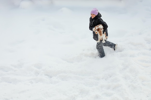 Ragazza giovane, donna si fa strada attraverso cumuli di neve, porta in braccio il cane Cavalier King Charles Spaniel. Vestiti caldi, giornata invernale innevata, felicità, gioia.