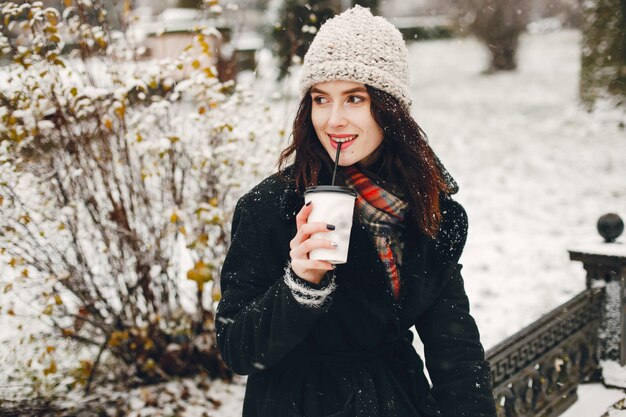 ragazza giovane ed elegante in cappotto nero e cappello bianco bere caffè in città invernale